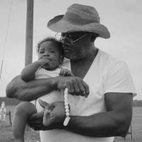 A monochrome image of an older man wearing a cowboy hat and white t-shirt holding a chubby baby.