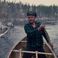 A man wearing a green plaid shirt and a hat rows a boat down a river surrounded by a snowy forest.
