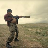 A woman wearing a knit hat and plaid flannel shirt with an exposed pregnant belly shooting a machine gun in an empty field.
