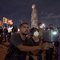 A man and a boy hold a camera at a protest. There is a crowd of people holding flags and a tall statue of a person in the background. The boy is wearing a white mask over his mouth. 