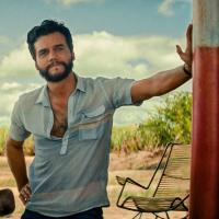 A man with a beard wearing a blue shirt leaning with one hand against a pole at what looks like a gas station. There is a yellow car to his right and it appears to be an arrid climate.