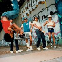 A group of people dancing in front of a wall covered in bold graffiti. One of the people in the air doing a backflip and another is holding a large boom box.
