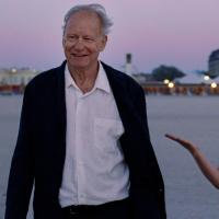 An older man wearing a suit walks on a beach at dusk beside a young blonde woman wearing a sparkly silver dress who is gesturing joyously.
