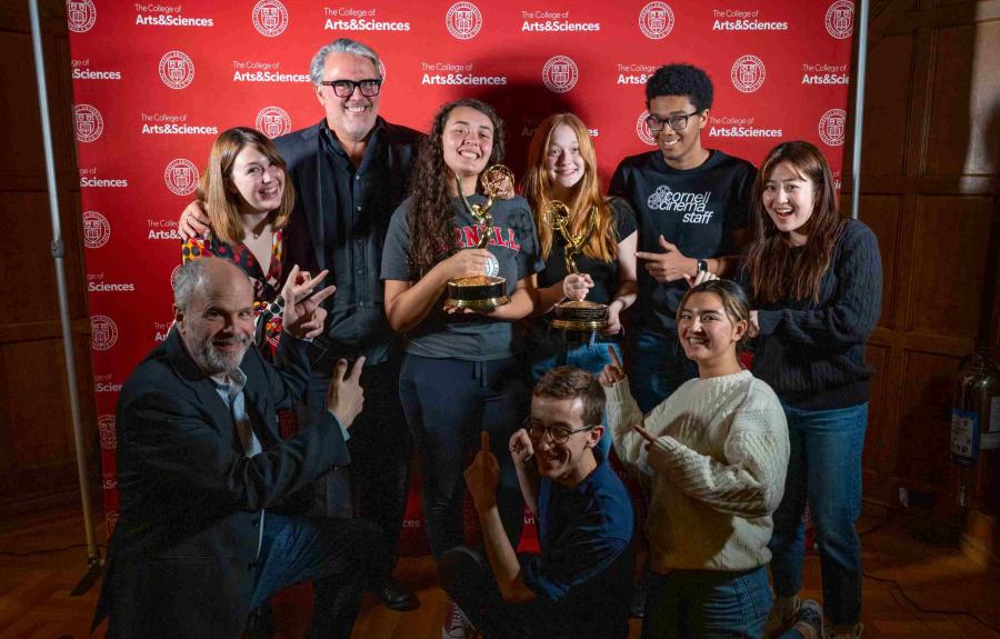 A group of people standing in front of a red backdrop and gathered around a woman who is holding a gold Emmy Award.