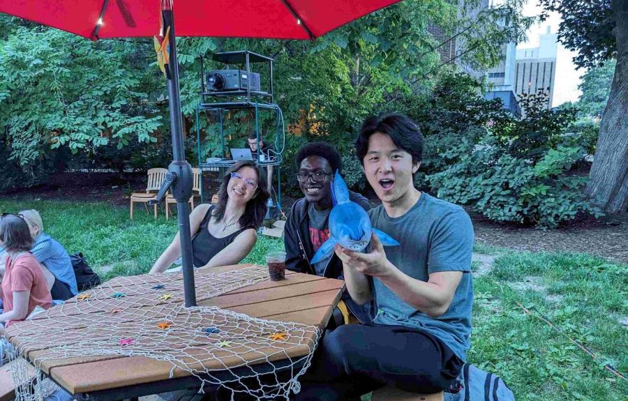 Three students sitting at picnic table. One student is holding a mini blow-up shark.