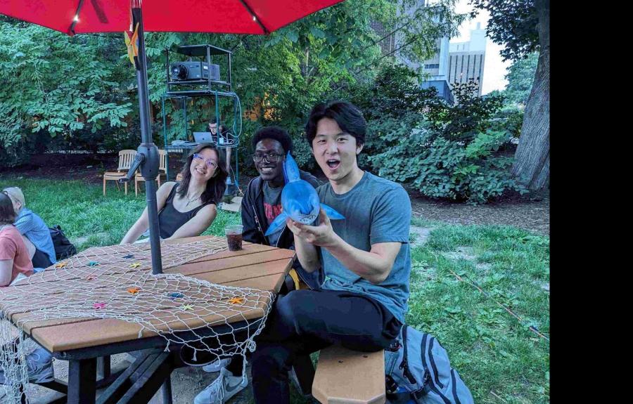 Three students sitting at picnic table. One student is holding a mini blow-up shark.