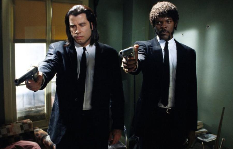 Two men in suits stand side by side pointing hand guns.