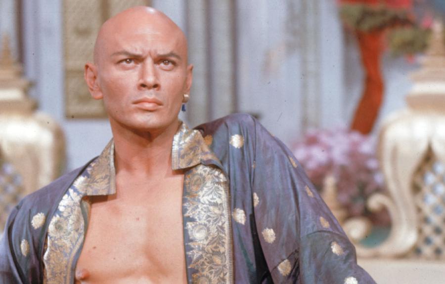 A scowling bald man wearing an open tunic with gold embroidery.