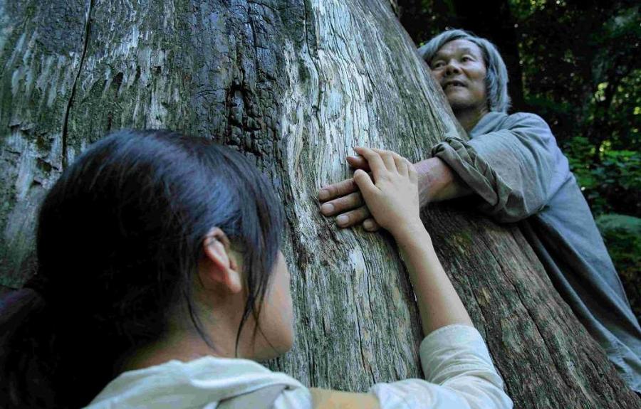 A young woman and an old woman hold hands while hugging a large tree.
