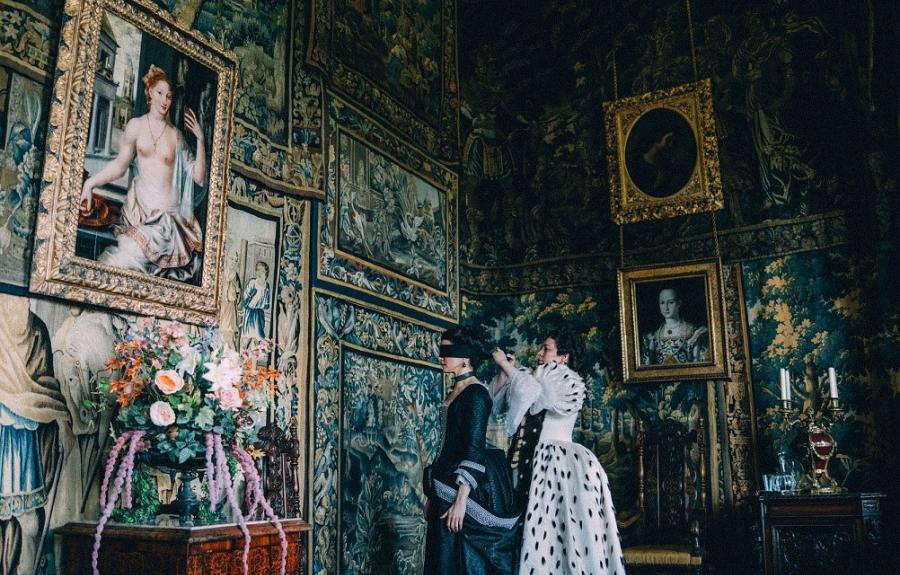 A woman in a white dress ties a blindfold over the eyes of a woman in a black dress inside a room decorated with ornate tapestries.