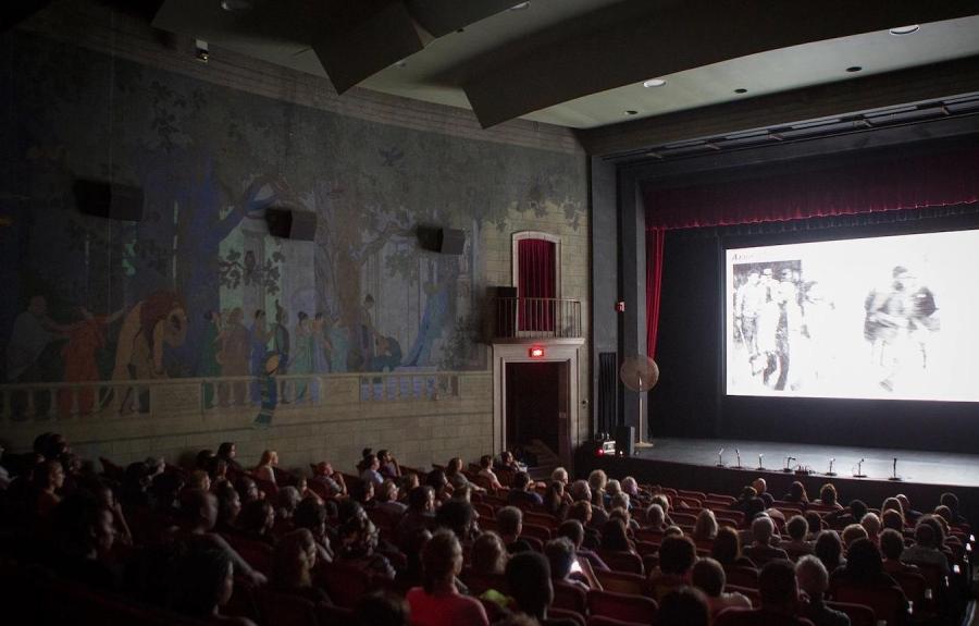An old movie theater filled with people with a black-and-white image on screen.
