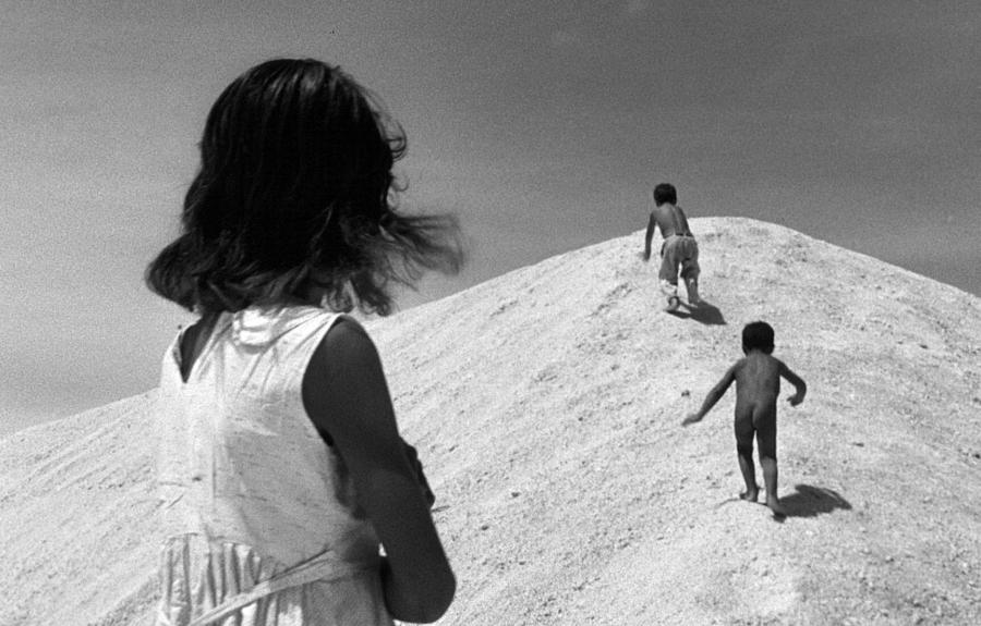 A black and white photo of two children climbing a pile of sand with an older child looking on.