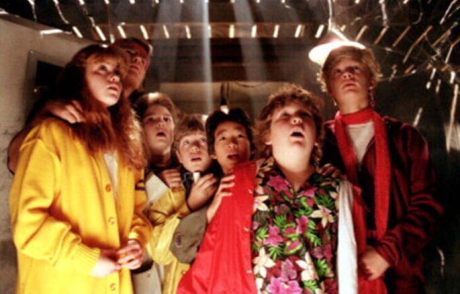 A group of children dress in colorful clothing standing in a basement beneath wooden floorboards.