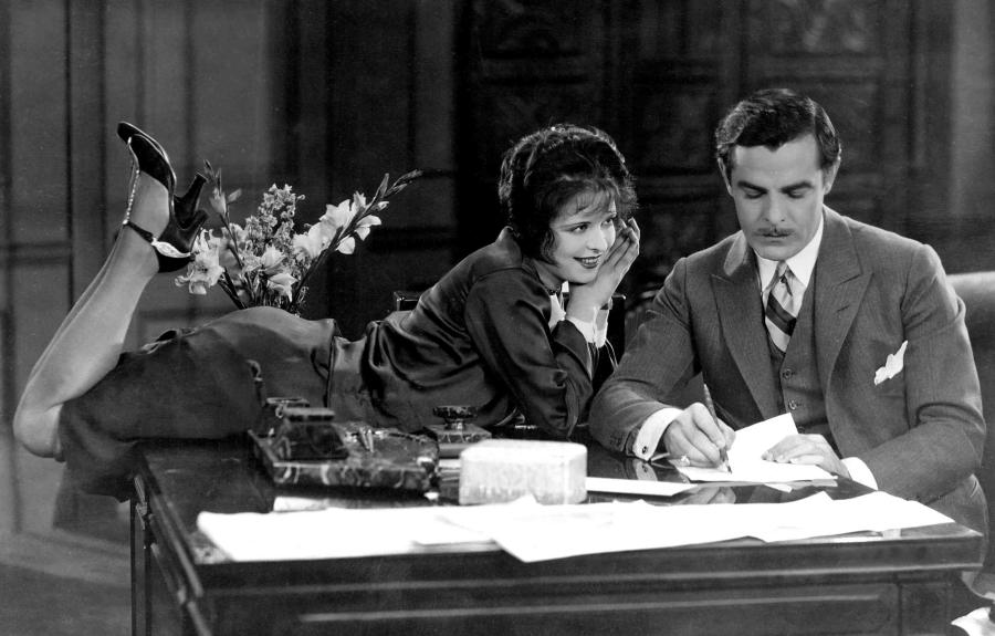 An old fashioned photograph of a women perched on a desk looking longing at a man who is ignoring her and writing on a piece of paper.