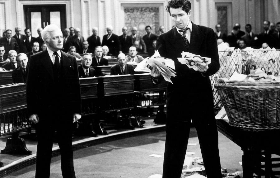 A forlorn looking man standing in the center of a courtroom holding a disorganized stack of papers in his hand.