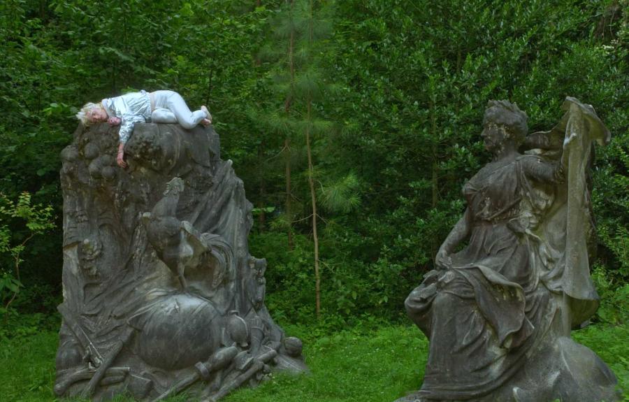 A man dressed in light blue sleeping atop a stone statue in a lush garden.