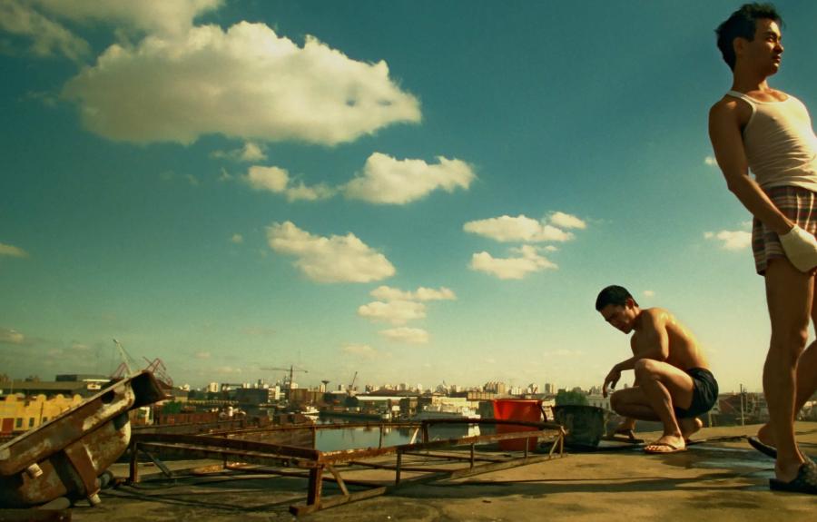 One man standing and one man crouching next to an industrial dock beneath a blue sky.