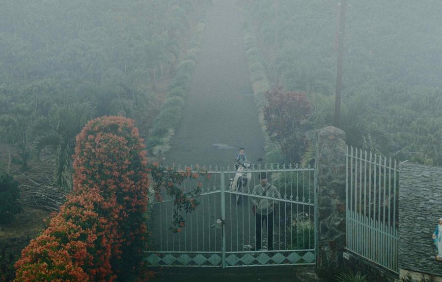 A man stands in front of an iron gate with a child sitting on a motorbike just behind him.