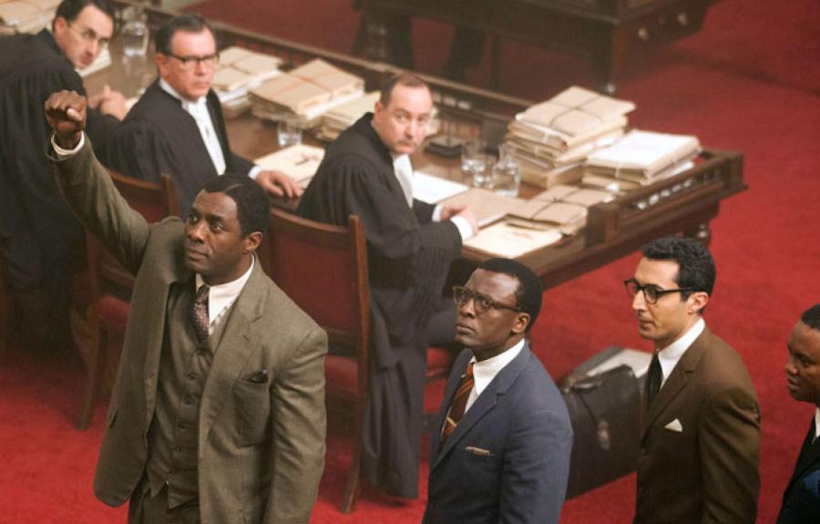 An aerial shot of a man in a suit raising his fist, while others look on in a courtroom with a red rug.