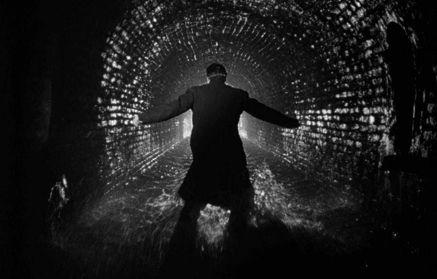 A monochrome image of a man running inside a dark tunnel with brick walls.