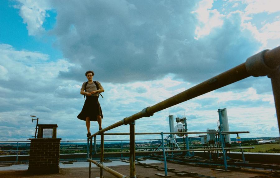 Person standing on a metal railing on a rooftop, with an industrial area and cloudy sky in the background
