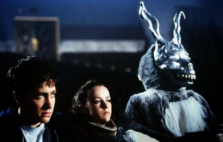 Three individuals sit in a dimly lit setting; one wears a large rabbit costume with sharp teeth and prominent ears