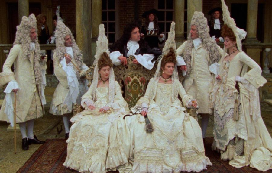Group of people in ornate 17th or 18th-century costumes with wigs, posed in front of a columned building
