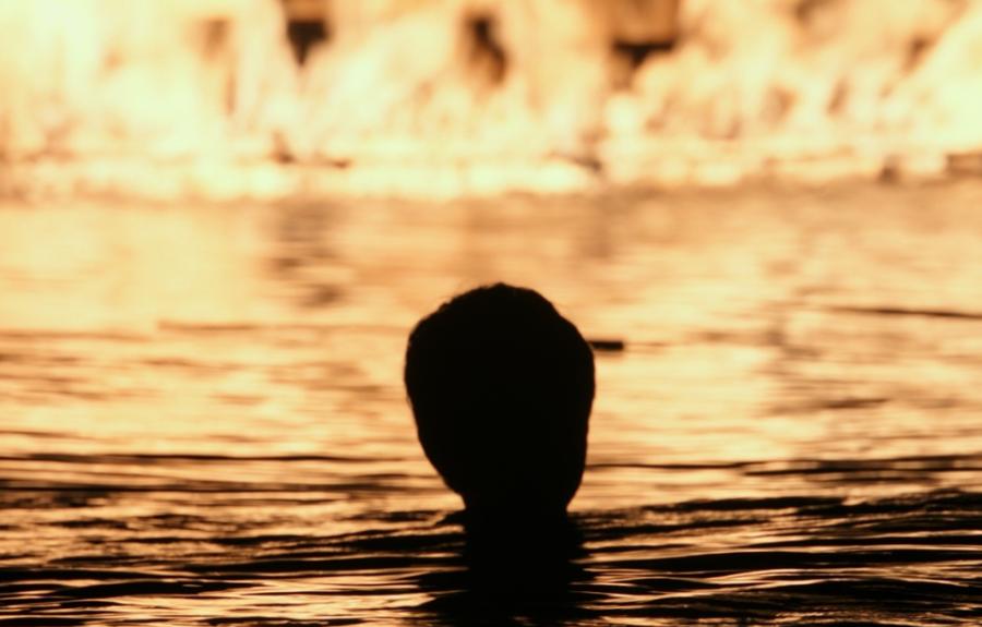 A shadowy image of a boy in the water looking at a boat on fire.