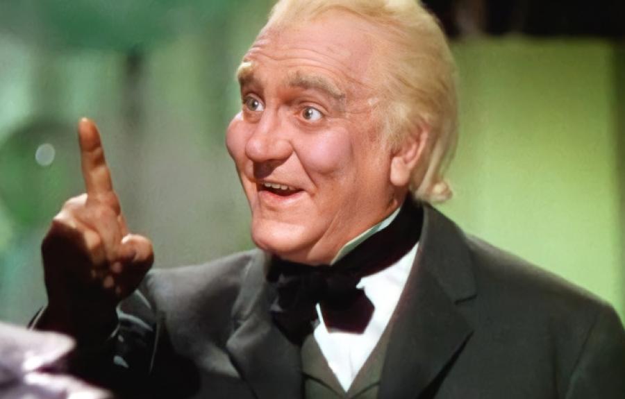 A cheerful man with blonde hair and wearing a dark green suit with a bow tie gestures with a pointed finger.
