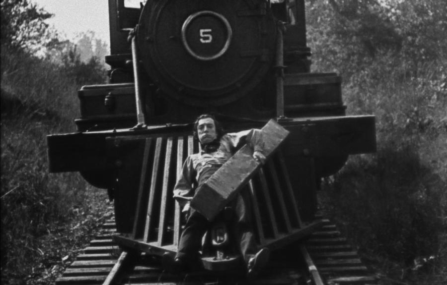 A black-and-white photo of a man clinging to the front of a steam engine train and holding a large block of wood.