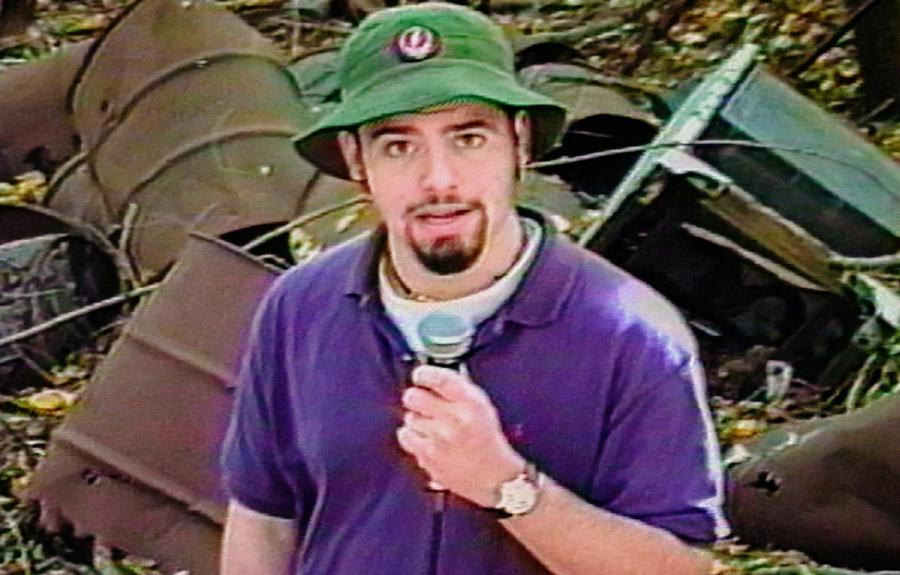 A person in a green hat and purple shirt holds a microphone in front of rusted metal barrels and debris.