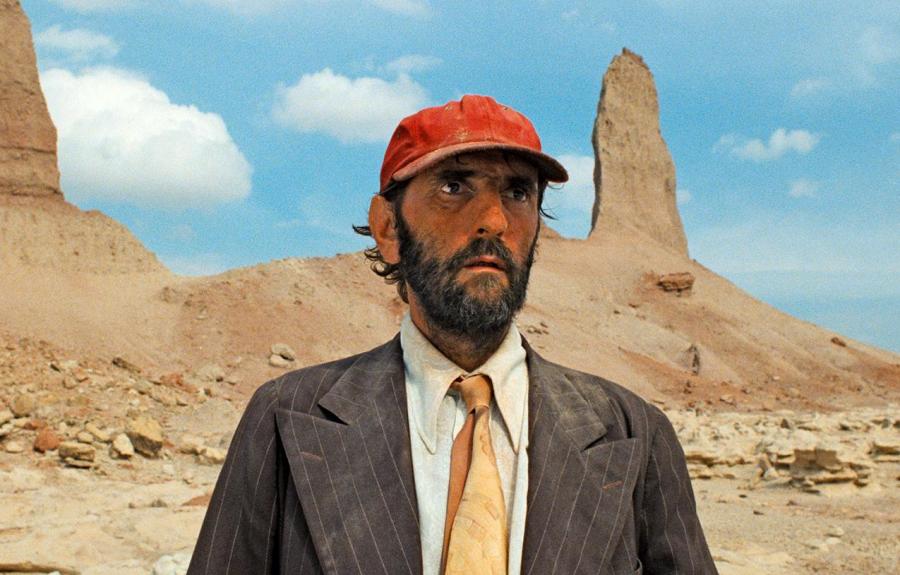 A person in a red cap, suit jacket, and orange tie stands in a desert landscape with rock formations in the background.