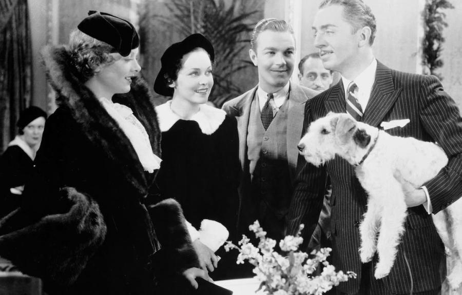 Black and white photo of four people in formal attire; the man on the right holds a small dog