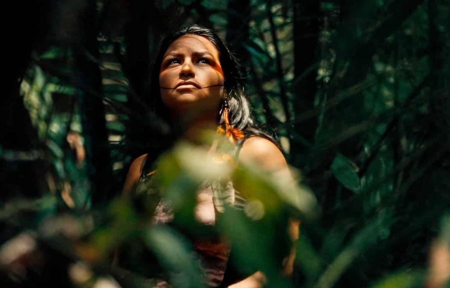 Person standing in a dense forest with lush green foliage; their face is partially covered by leaves and branches