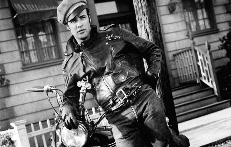 A man with a leather jacket and military cap leaning casually against a motorcycle.