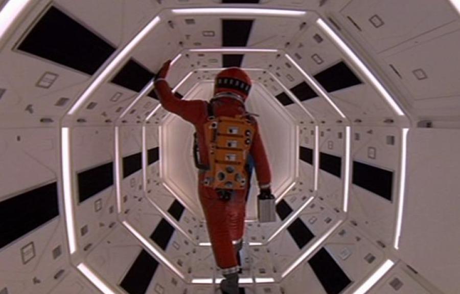 A person in a red space suit walks through a brightly lit, hexagonal corridor with futuristic design.