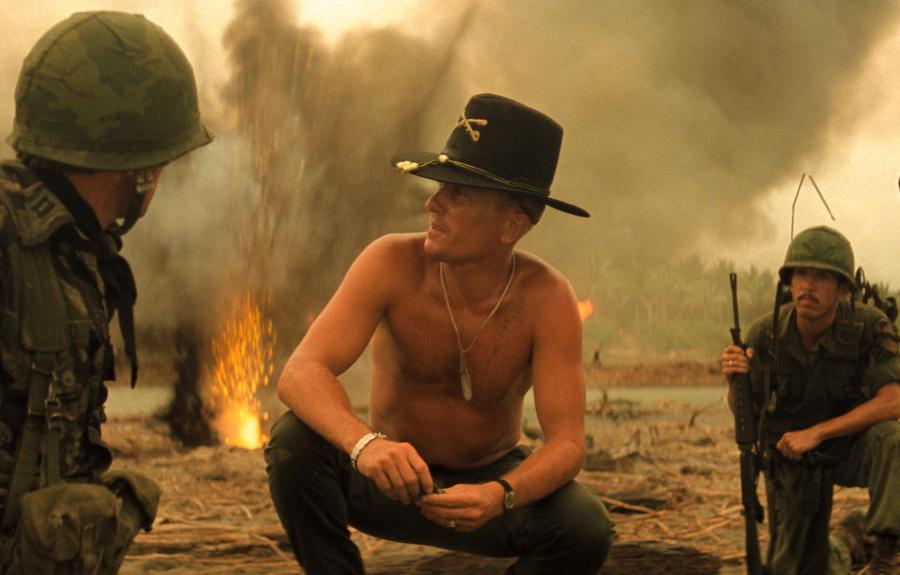 Soldiers gather in a smoky war zone with fire in the background, suggesting recent combat activity. A shirtless man in a cavalry hat speaks to another soldier, while others stand nearby in military gear.