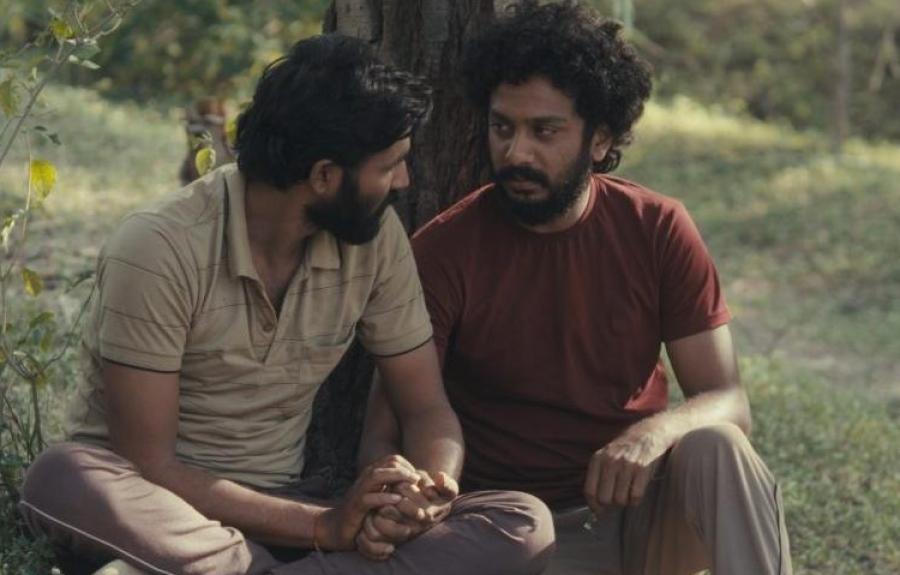 Two people sit on the ground outdoors, leaning against a tree and holding hands. The natural setting features grass and sparse vegetation, creating a quiet moment of connection.