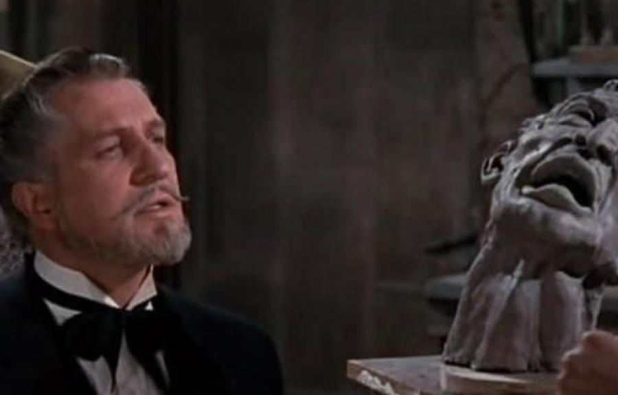 A man in a black suit and bow tie sits beside a dramatic sculpted bust with an anguished expression. The indoor setting includes shelves and tools, suggesting an artist’s studio or workspace.