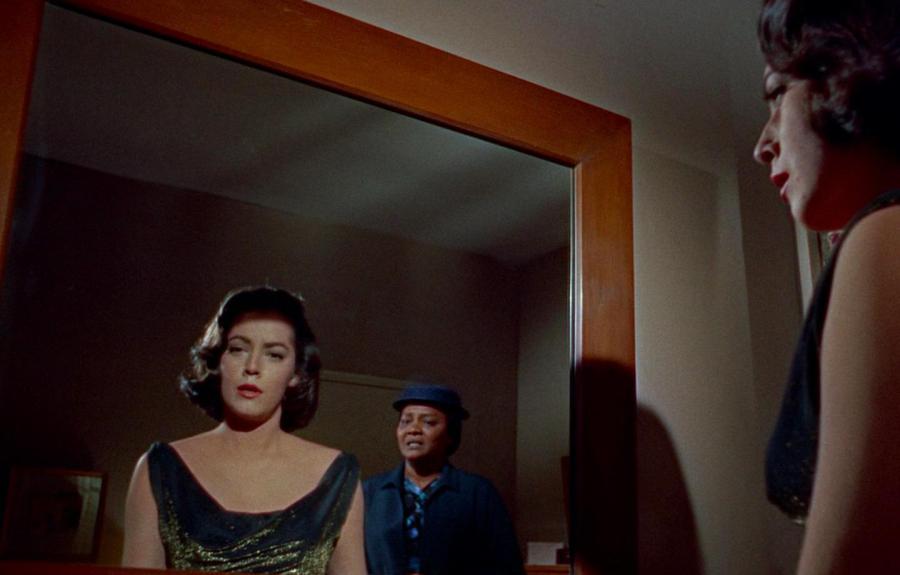 A woman in a black dress with a shimmering neckline looks into a mirror, revealing her reflection and a second person in a blue outfit and hat standing behind her. 