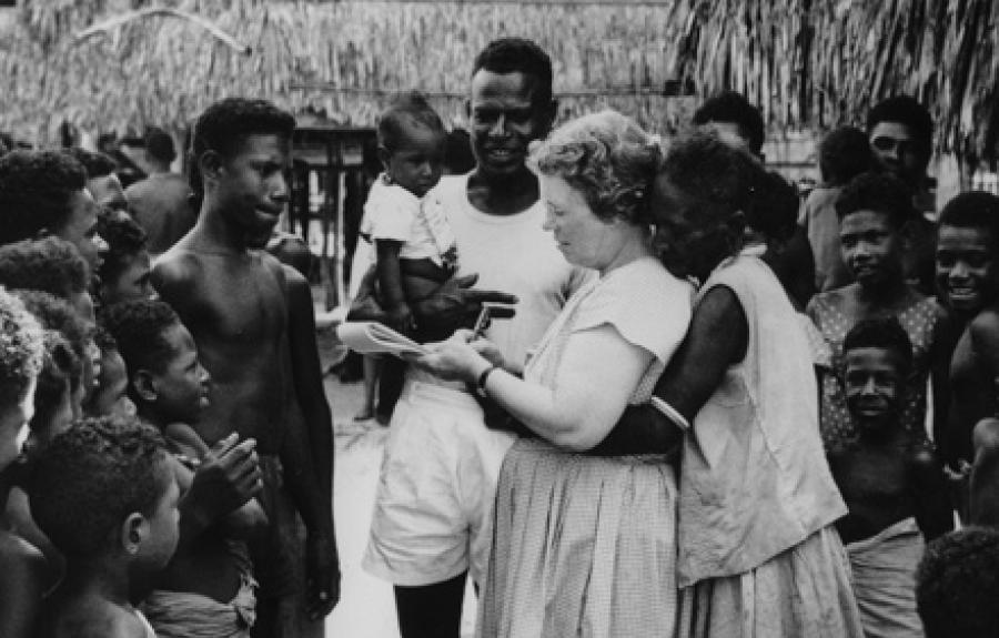A woman writes on a note pad encircled by a group of men, women, and children. Another woman embraces her from behind.