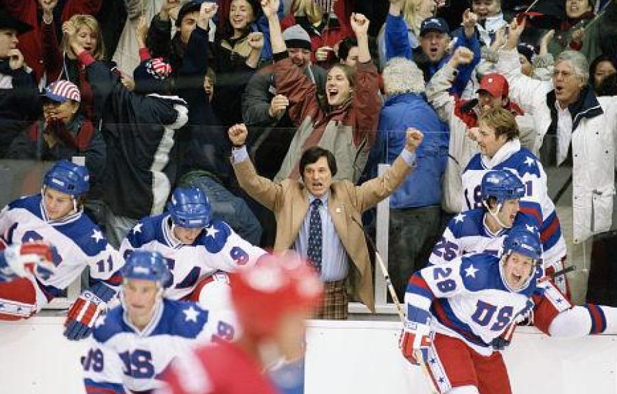 Players in white USA hockey jerseys cheer from the bench as fans erupt behind them. A man in a tan jacket and tie raises his fists in celebration. The scene captures a moment of triumph and national pride during an intense game.