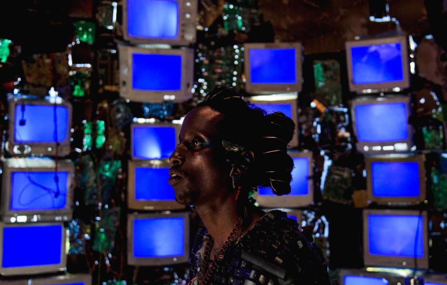 A person stands in front of a wall of old CRT monitors, each displaying a solid blue screen. They wear elaborate accessories made from electronic components, with coils in their hair and circuit boards on their clothing. The background is filled with wire