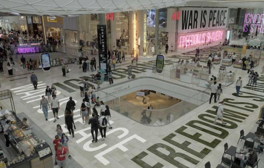 A busy shopping mall features retail stores like H&M and John Lewis, with crowds walking and dining. Bold neon slogans—“WAR IS PEACE,” “FREEDOM IS SLAVERY,” and “IGNORANCE IS STRENGTH”—are prominently displayed, evoking Orwellian themes amid the consumeri