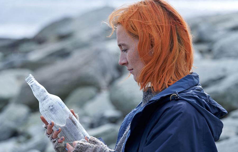 A woman with bright orange hair stares at a sand-filled glass bottle on a cold beach.