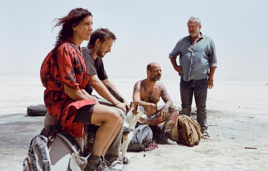 Four people and a dog gather in a desert landscape. Three sit on the ground or on bags—one in a red and black patterned outfit, one in a black shirt, and one shirtless with tattoos and colorful pants. A fourth person stands nearby in a blue shirt and jeans. Backpacks and gear surround them, and the clear sky emphasizes the stark, sandy terrain.