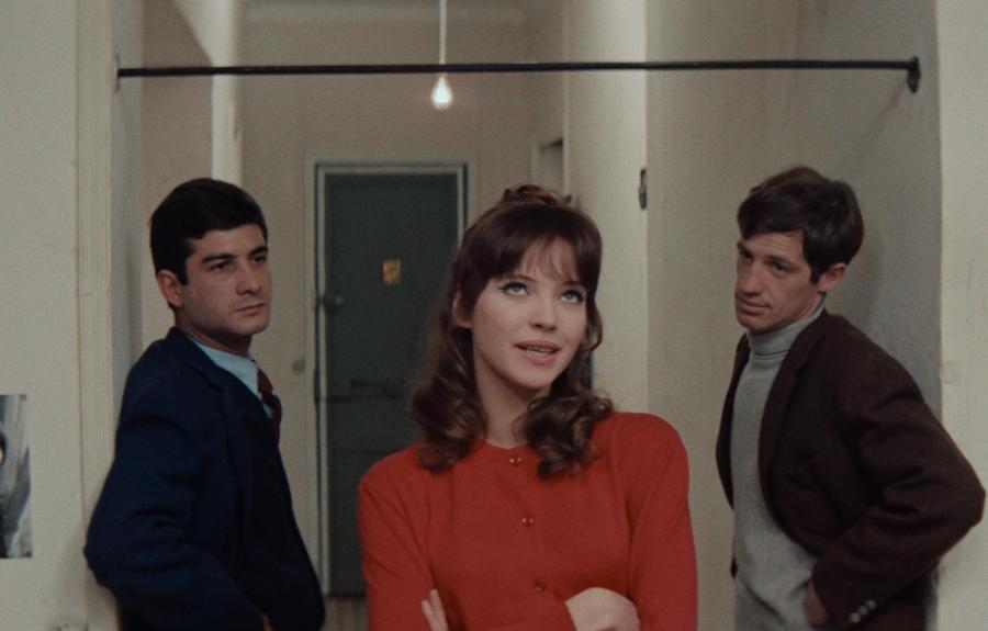 Three people stand in a hallway with white walls and a green door, engaged in a tense conversation. The central figure in red gestures while the others listen.