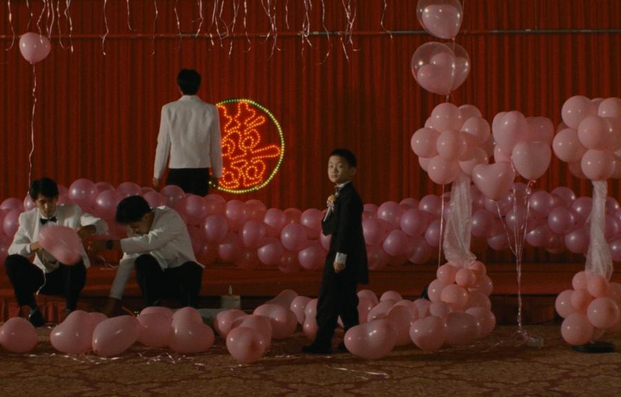 A decorated event space with pink balloons arranged in clusters and scattered on the floor, featuring people in formal attire. A red backdrop with a circular neon design is visible behind the stage area.