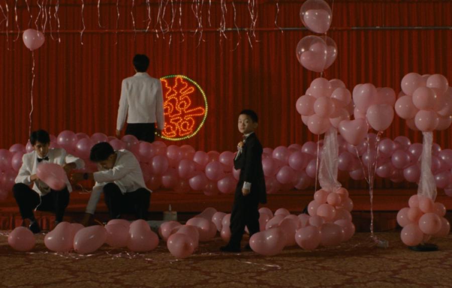 A decorated event space with pink balloons arranged in clusters and scattered on the floor, featuring people in formal attire. A red backdrop with a circular neon design is visible behind the stage area.