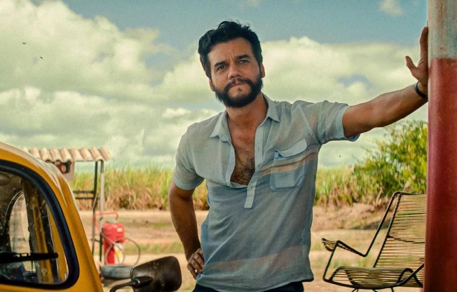 A man with a beard wearing a blue shirt leaning with one hand against a pole at what looks like a gas station. There is a yellow car to his right and it appears to be an arrid climate.